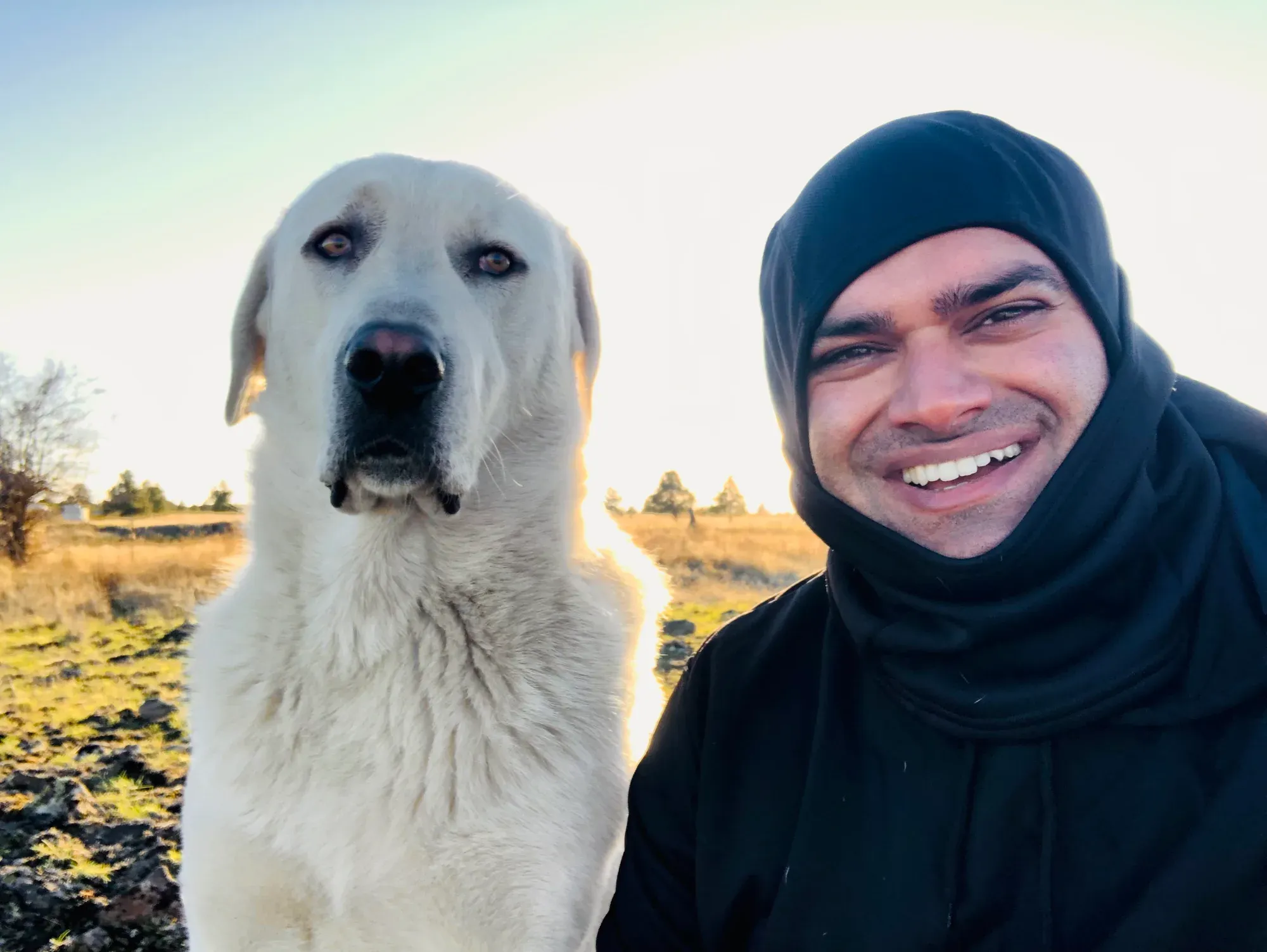 Himanshu Mehru smiling next to Lincoln, a large white livestock guardian dog, on the open prairie of his Creston homestead at golden hour