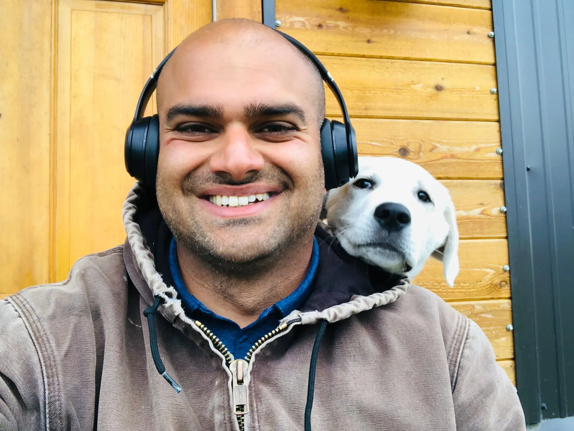 Himanshu Mehru wearing headphones and smiling while a white puppy rests its head on his shoulder